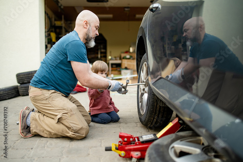 Cute toddler boy helping his father to change car wheels at their backyard. Father teaching his little son to use tools.