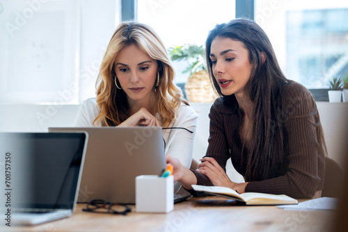 Two elegant attractive businesswomen talking while working together with laptop in a modern startup