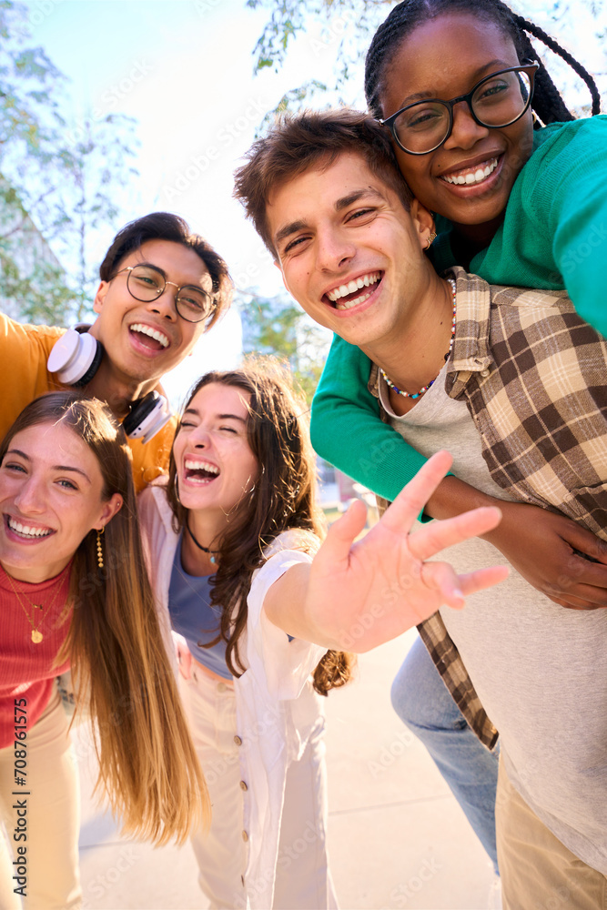 Obraz premium Vertical multiracial group of teenagers taking a selfie looking front camera laughing and having fun piggybacking together outdoor. Young students enjoying their free time with friends in street city