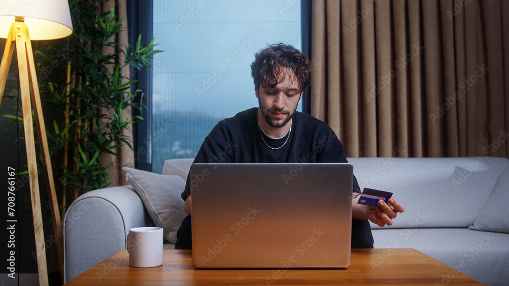 Curly hair male enter credit card number on laptop shopping online, makes secure easy distant payment while sitting on sofa at home.	
