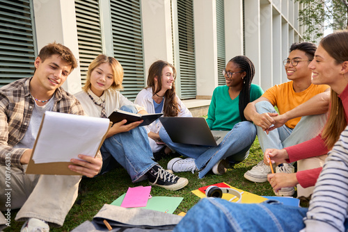 Group of young international university students sitting on the grass outside the faculty building. Friends gathered happily studying together with workbooks and laptop on campus