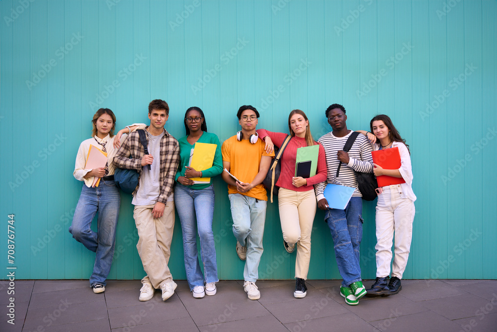 Obraz premium Serious international students posing standing looking at the camera. Young people multicultural friends concentrated and embracing with backpacks and workbooks in a blue wall background