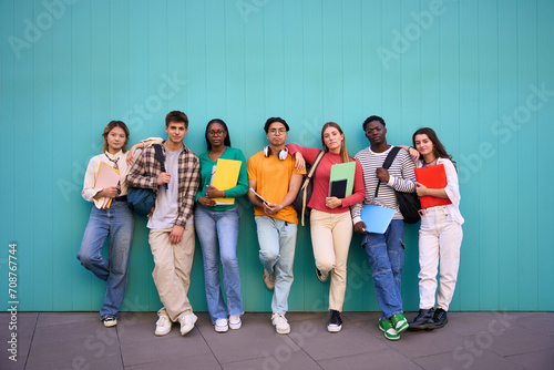 Serious international students posing standing looking at the camera. Young people multicultural friends concentrated and embracing with backpacks and workbooks in a blue wall background photo