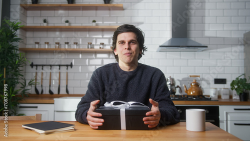 Young vlogger man sitting in kitchen, showing gift box, advertising product , talking to camera, recording video 
