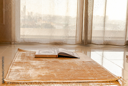 A muslim carpet set on the floor, beige brownish color with a mosque, curves and rectangle patterns. 
An opened Holy coran book placed on the rug in front of a balcony window.  photo