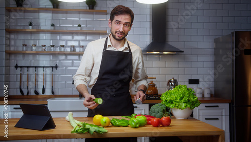 Bearded young vlogger man chef in an apron recording video cooking lesson about vegetarian meal in modern kitchen 