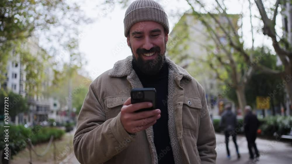 Front view of smiling young Caucasian man using smartphone while walking in city street. Male having fun looking his mobile through a urban area on a winter christmas day. 