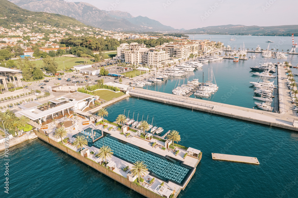 Long swimming pool on the wide pier of a luxury marina with moored yachts and modern houses on the shore