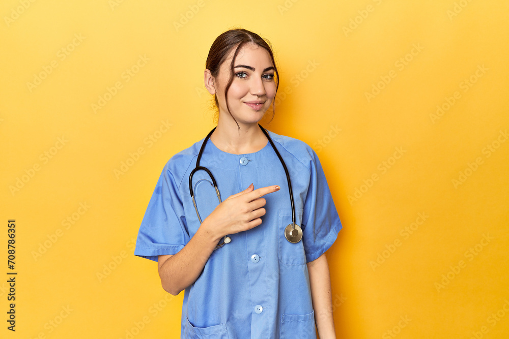 Young nurse in a yellow studio smiling and pointing aside, showing something at blank space.