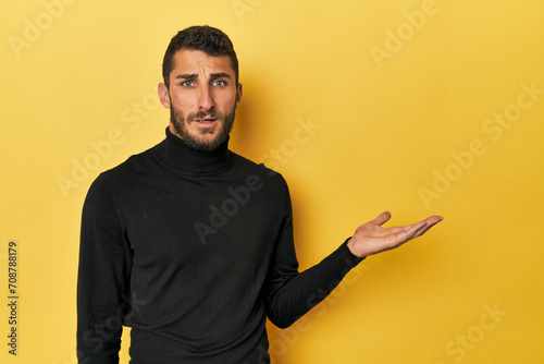 Young Hispanic man on yellow background impressed holding copy space on palm. photo