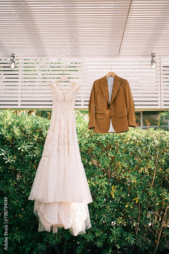 Groom suit and bride dress hang on hangers on a wooden canopy in the garden © Nadtochiy