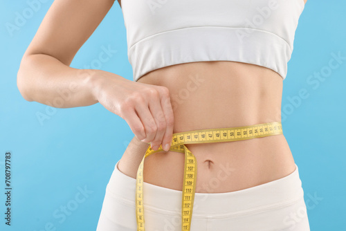 Slim woman measuring waist with tape on light blue background, closeup. Weight loss photo