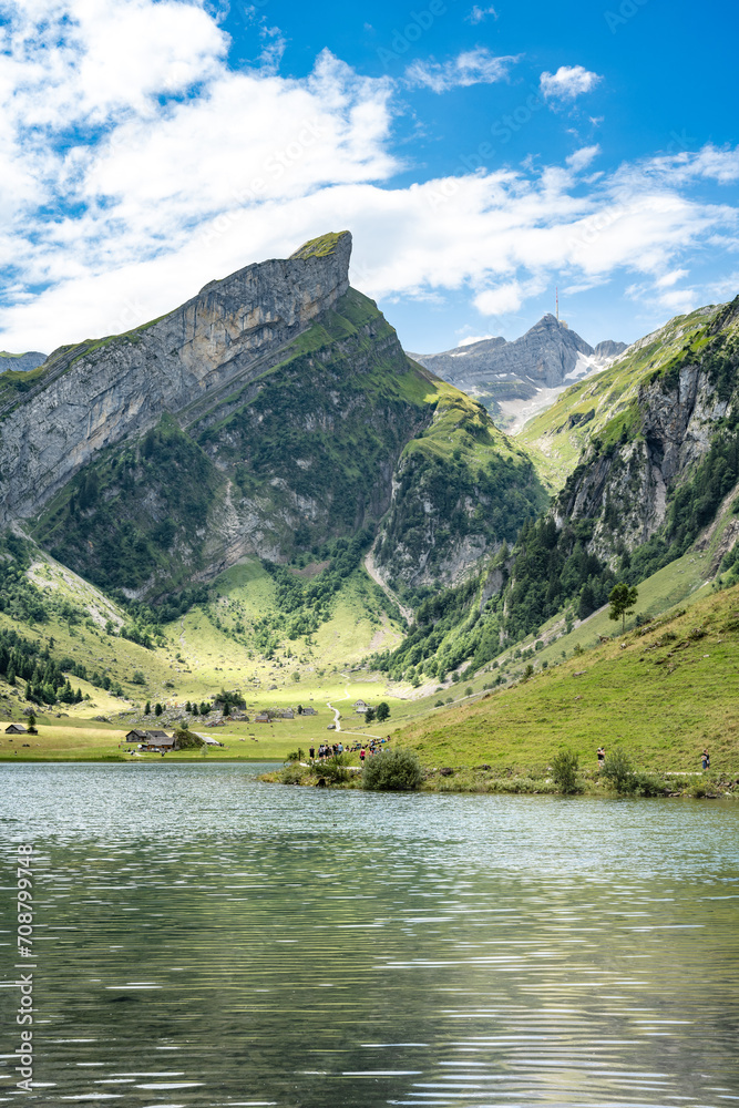 View of tourists walking along an alpine lake in a green valley with a mountain peak in the background on a sunny day. Seealpsee, Säntis, Wasserauen, Appenzell, Switzerland.