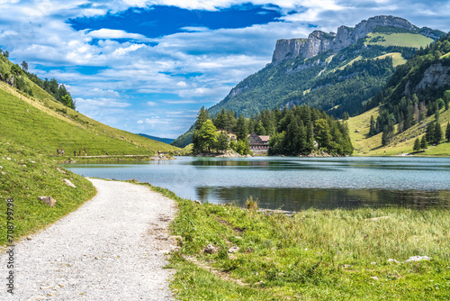 Panoramic view of tourists walking along an alpine lake in a green valley with a mountain huts in the background on a sunny day. Seealpsee, Säntis, Wasserauen, Appenzell, Switzerland.
