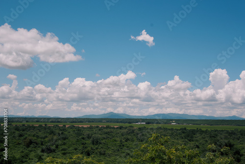 Expansive view of lush greenery, flat fields, distant mountains, and a vast sky dotted with fluffy clouds.