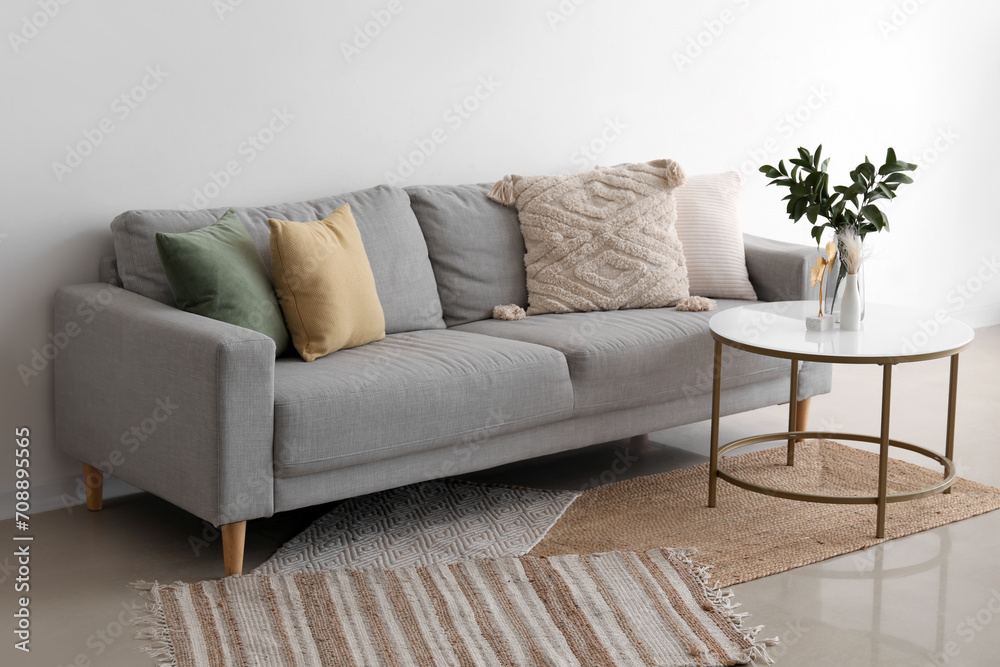 Cozy grey sofa with cushions and plant branches in vase on table near light wall Cozy grey sofa with cushions and plant branches in vase on table near light wall