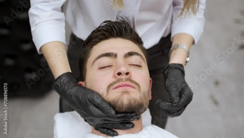 Barber gives relaxing neck face massage to male client lying in chair with eyes closed. Professional wears black gloves for skincare routine in barbershop setting, enhancing client comfort. photo
