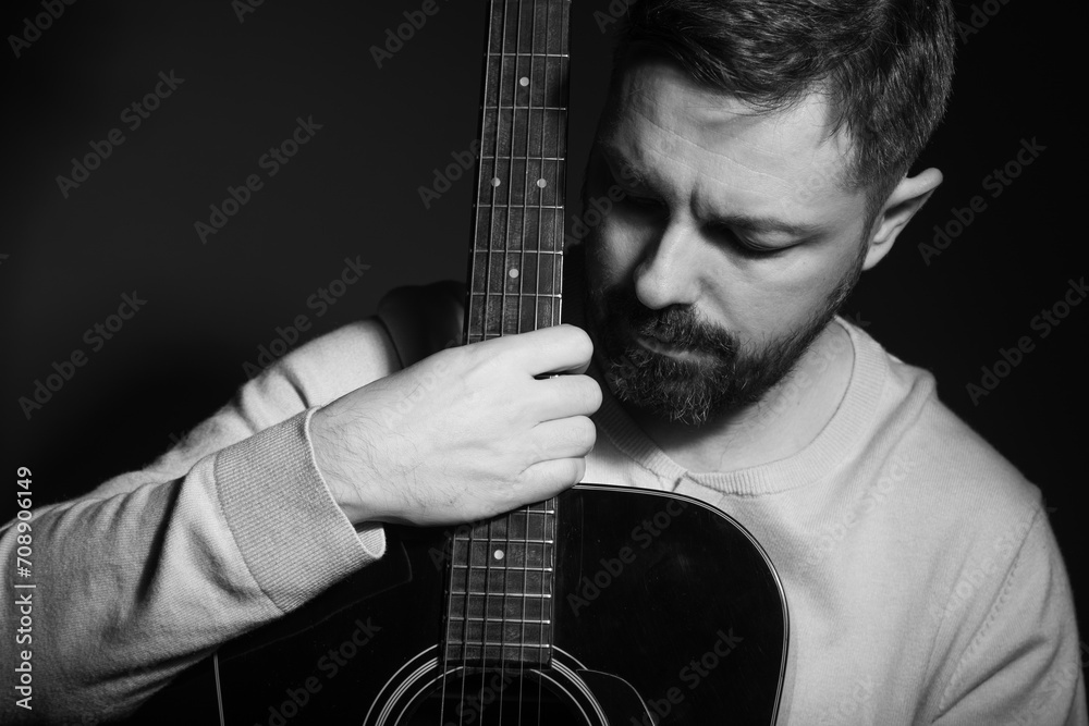 Black and white portrait of a caucasian man holding a guitar in his lap. He is in his 40s and is wearing a white sweater. He has brown hair and a beard. The photo was taken in a studio. - obrazy, fototapety, plakaty 