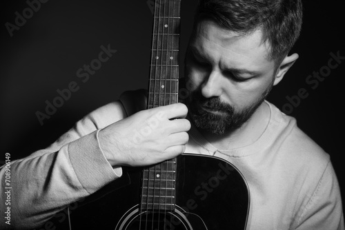 Black and white portrait of a caucasian man holding a guitar in his lap. He is in his 40s and is wearing a white sweater. He has brown hair and a beard. The photo was taken in a studio. photo