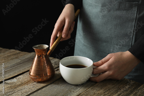 Turkish coffee. Woman with cezve and cup of freshly brewed beverage at wooden table against black background, closeup