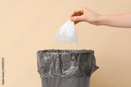Female hand throwing used tissue into trash bin near beige wall, closeup photo