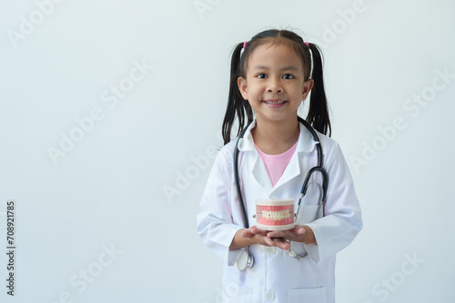 Young Asian girl wearing a doctor's uniform hangs a stethoscope around her neck in a hand holding a dental model with a smiling face on a white background. Medical health care concept.