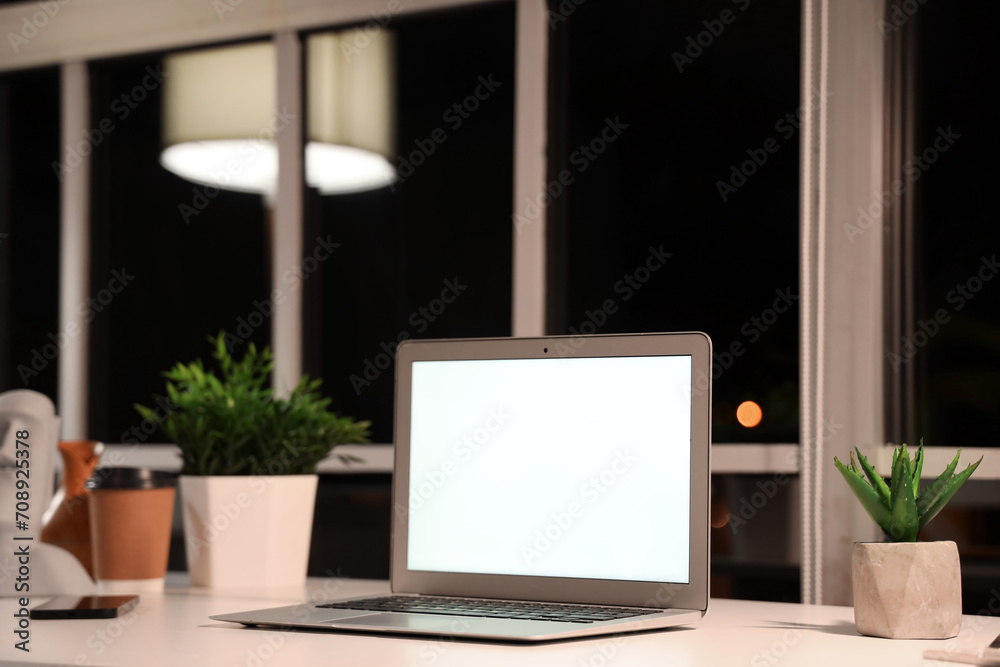 Fototapeta premium Blank laptop and plants on desk in office at night, closeup