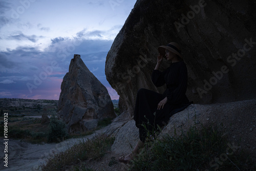 Cappadocia. An Asian girl in a black dress and a wide-brimmed hat is photographed against the backdrop of mountains at dusk.
