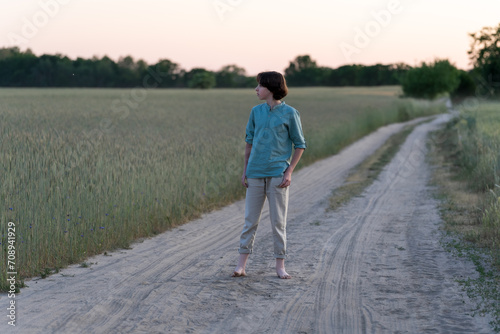 A barefoot teenager in comfortable clothes stands on a dirt road. © Sergey Kohl