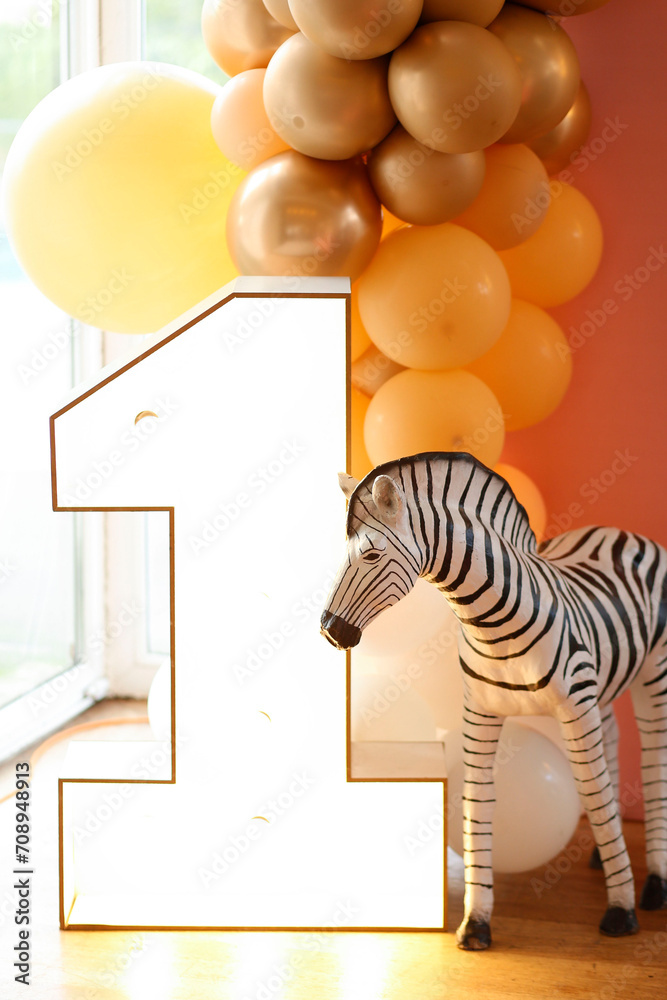 A playful and colorful first birthday party decoration featuring a large wooden number one, a zebra, and a balloon arch in shades of yellow and gold