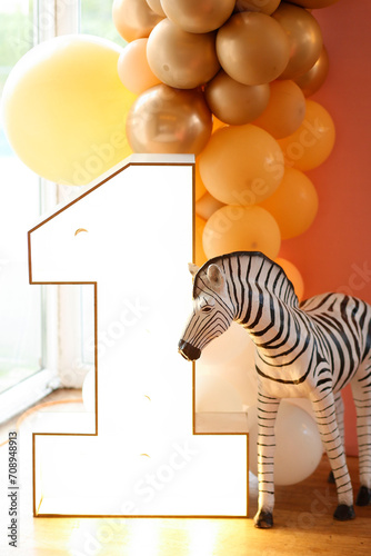 A playful and colorful first birthday party decoration featuring a large wooden number one, a zebra, and a balloon arch in shades of yellow and gold