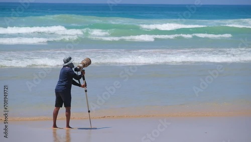 A photographer on a beautiful beach takes pictures of surfers with a camera with a large telephoto lens and a monopod.