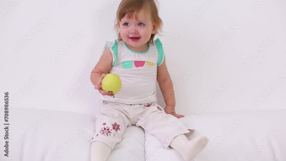 Cute little girl sits on back of white couch and holds apple Cute little girl sits on back of white couch and holds apple