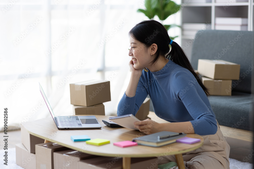 Asian woman starting a small business at home working with parcel boxes and calculating revenue and profit from sales. Asian woman starting a small business at home working with parcel boxes and calculating revenue and profit from sales.