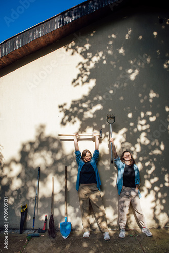 Two happy lesbian girls pose and laugh with garden tools near the wall in the shade of trees and the rays of the sun © Kate Stock
