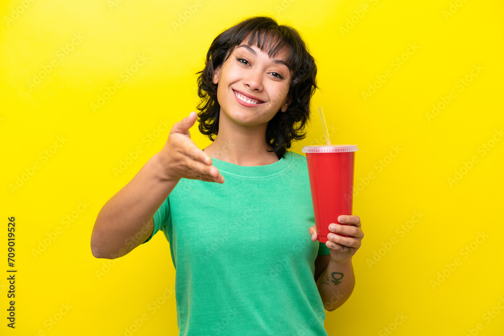 Young Argentinian woman holding a soda isolated on yellow background shaking hands for closing a good deal Young Argentinian woman holding a soda isolated on yellow background shaking hands for closing a good deal