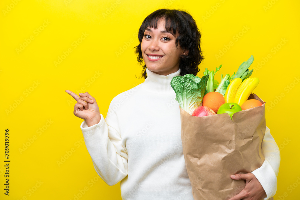 Young Argentinian woman holding a grocery shopping bag isolated on yellow background pointing finger to the side Young Argentinian woman holding a grocery shopping bag isolated on yellow background pointing finger to the side