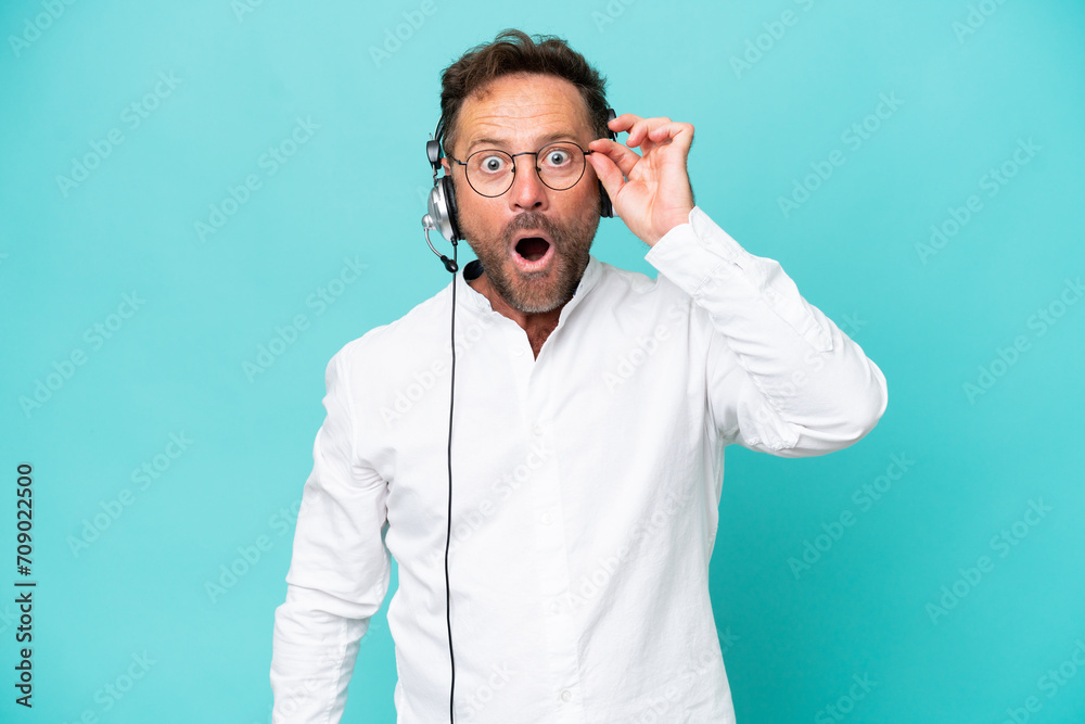 Telemarketer caucasian man working with a headset isolated on blue background with glasses and surprised Telemarketer caucasian man working with a headset isolated on blue background with glasses and surprised
