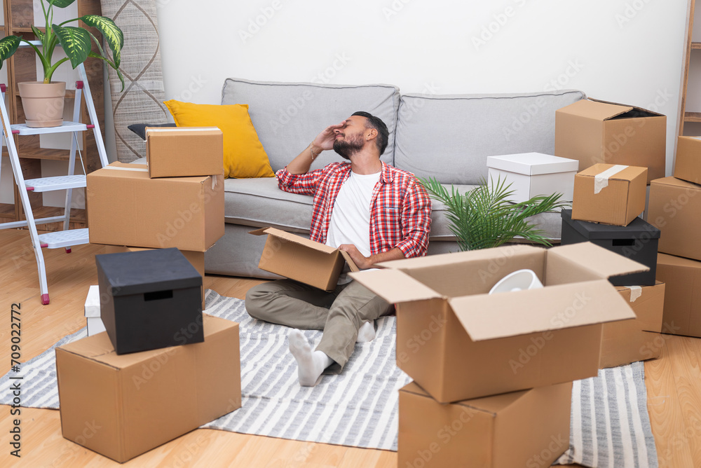 Smiling man in shirt with delighted expression sits on floor with carton box enjoying moving to spacious apartment with preparing for arrangement of modern house Smiling man in shirt with delighted expression sits on floor with carton box enjoying moving to spacious apartment with preparing for arrangement of modern house