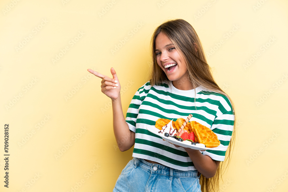 Young caucasian woman holding waffles isolated on yellow background pointing finger to the side and presenting a product Young caucasian woman holding waffles isolated on yellow background pointing finger to the side and presenting a product