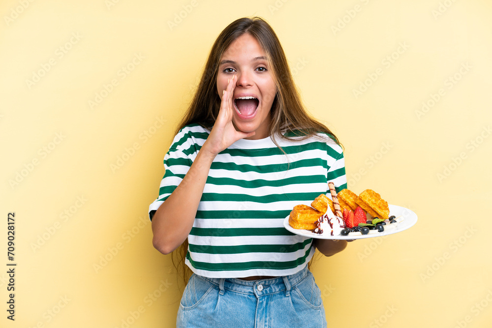 Young caucasian woman holding waffles isolated on yellow background shouting with mouth wide open Young caucasian woman holding waffles isolated on yellow background shouting with mouth wide open