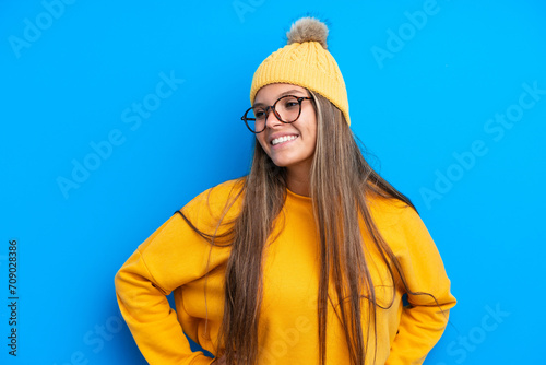 Young caucasian woman wearing winter clothes isolated on blue background posing with arms at hip and smiling