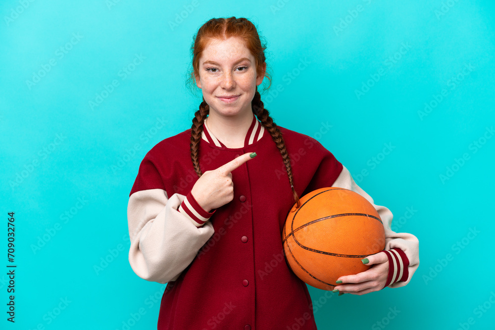 Young reddish woman playing basketball isolated on blue background pointing to the side to present a product