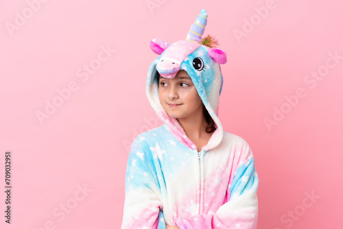Little caucasian woman wearing a unicorn pajama isolated on pink background looking to the side and smiling