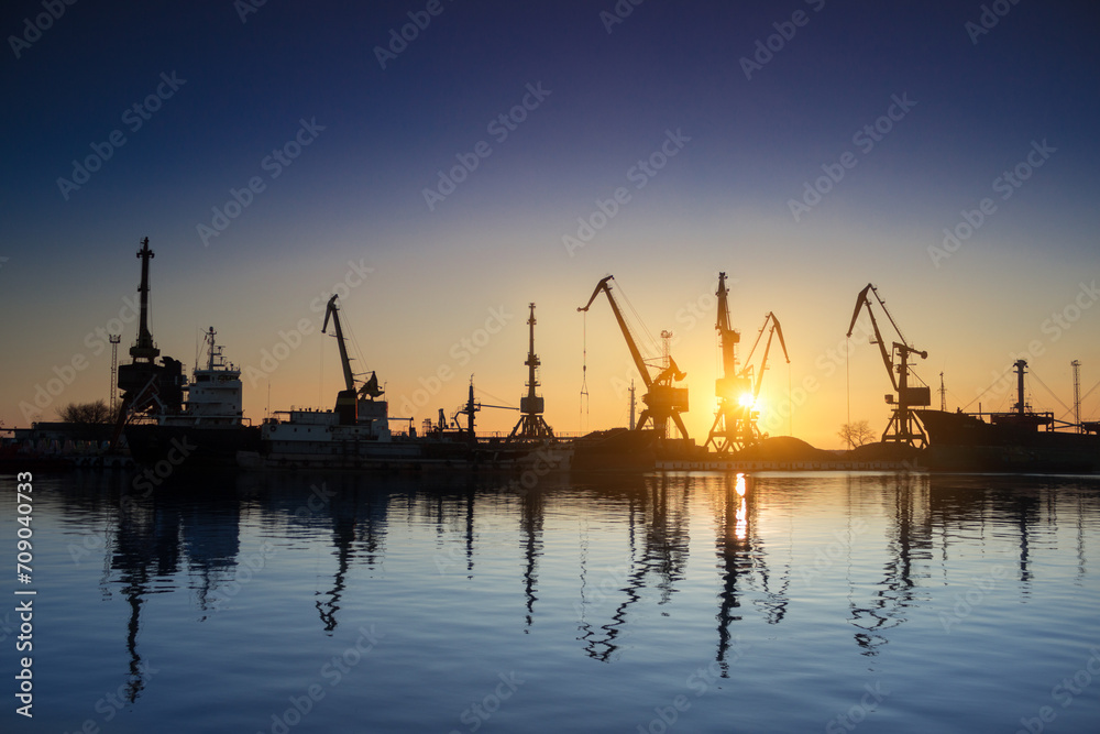Sunset silhouettes cargo ships, cranes at sea port. Industrial loads freight at dock. Trade, transport infrastructure. Maritime logistics, global shipping. Clear sky reflects calm water.