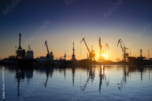 Sunset silhouettes cargo ships, cranes at sea port. Industrial loads freight at dock. Trade, transport infrastructure. Maritime logistics, global shipping. Clear sky reflects calm water.