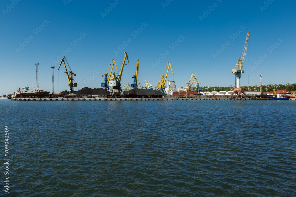 Derelict port cranes line harbor, symbolizing industrial decline. Unused loading equipment stands against clear sky, hinting at past maritime commerce vitality, awaiting repurpose or dismantle.