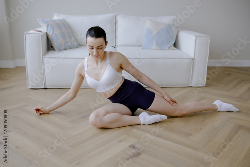 Young woman in sportswear doing exercises on the floor at home. Beautiful European appearance. There is a white sofa in the background. Muscle stretching. Morning work-out.