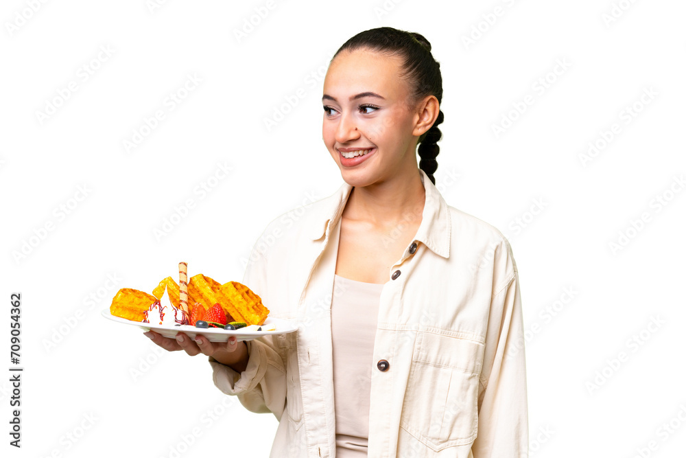 Young Arab woman holding waffles over isolated background looking to the side and smiling Young Arab woman holding waffles over isolated background looking to the side and smiling