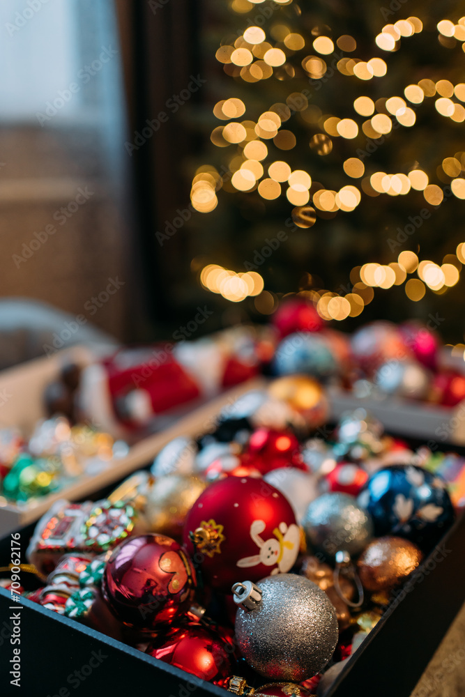 A box with Christmas toys. In the background there is an artificial Christmas tree with a garland with yellow lights, but without toys. Decorate the Christmas tree for the New Year